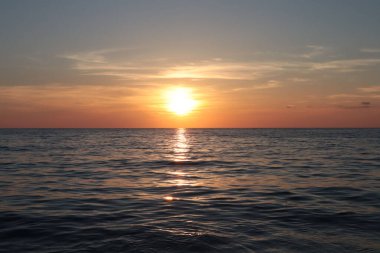 Denizde gün batımı, ufukta turuncu gökyüzü ve güneş, su yüzeyinde güneşin yansıması.