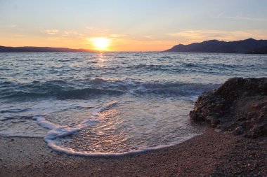 Sunset at sea, orange sky and clouds, sun and silhouette of mountains on the horizon, reflection of sun on water surface, on wet stones.  Croatia, Dalmatia, Baska Voda