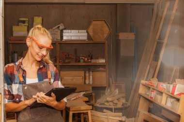 Mobilya fabrikasında güvenlik gözlüğü takan kadın marangoz portresi. Kereste ve marangozluk aletleri için bir depoda gülümsüyor..