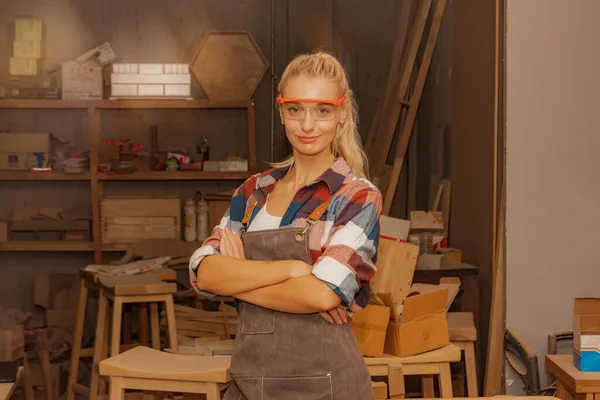 Portrait woman carpenter wearing safety glasses in a furniture manufacturing plant stands in a woodworking workshop holding workshop proudly looking at a smart camera, hands crossed arms.