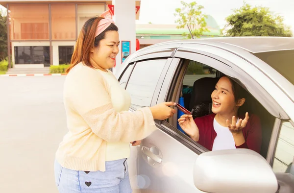 Fat girl friend happy to lend credit card to pay for fuel instead of cash with pleasure in order to collect points for beautiful girl who drives her while she parks her gas station at modern station.