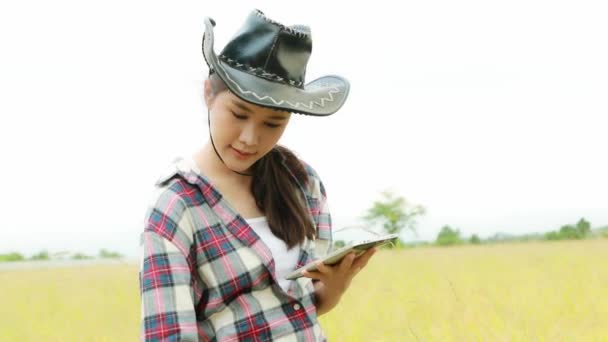 美丽的亚洲农民头戴礼帽 看着稻谷长在成熟的田里 在平板电脑上打字 在田里散步 快乐地收集着夏天的信息 农业综合企业 — 图库视频影像