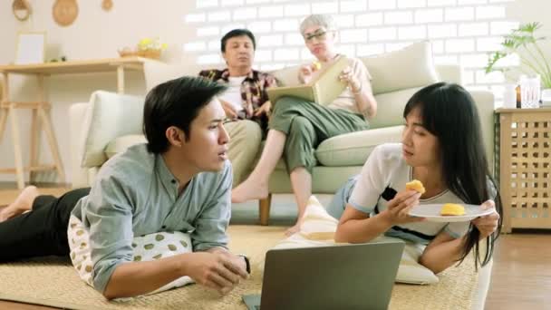 自宅のリビングルームで週末に集まるアジアの家族 妹はお菓子を食べることについて彼女をからかい 両親が禁止し 警告するまで彼女の兄と競争しました — ストック動画