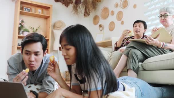 アジアの家族の週末は一緒に家のリビングルームで顔を取得暖かい家族の両親の息子と娘座っている間に軽食を持っているとラップトップとお母さんはお父さんとソファ読書に座って — ストック動画