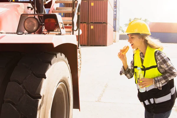 Female workers forklift drivers in the container loading yard eating hamburgers and drinking water breakfast before duty driving walking checking the safety of wheels and other parts for safety.