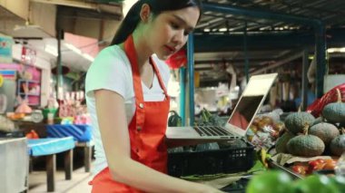 İş kadını internetten taze sebze pazarında dizüstü bilgisayar kullanarak pazarda modern ticaret sicili kullanarak müşterinin taze kırmızı biber siparişini arıyor ve taze sebze topluyor..