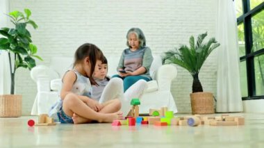 Evde torununa bakan nazik büyükannesi: Büyükannesi kanepede oturur ve torununun, erkek kardeşinin ve kız kardeşinin renkli tahta bloklarla oynayışını izler..