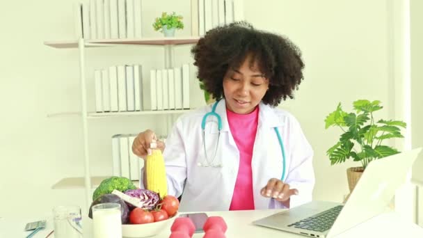 可爱的非洲裔美国女性营养学家笑着坐在桌子上 把有机蔬菜玉米放在网上 供病人在医院办公室的笔记本电脑上消费 — 图库视频影像