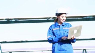 Happy smile field work of engineer, Portrait beautiful female electrician working solar panel station holding laptop patrolling outdoor hot weather, solar mounting structure with large solar panels.