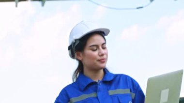 Happy smile field work of engineer, Portrait beautiful female electrician working solar panel station holding laptop patrolling outdoor hot weather, solar mounting structure with large solar panels.