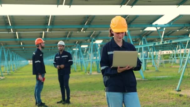 ハード帽子をかぶっている女性電気技師の労働者は 大きな太陽電池ステーションエリアで背景に男性エンジニアのチームと一緒にラップトップで太陽電池パネルを検査します — ストック動画