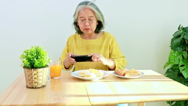 幸せな高齢者のアジアの女性は自宅で午後の軽食のためのダイニングテーブルに座って 彼女の携帯電話ギャラリーでケーキやパイのお土産を撮影するために彼女のスマートフォンを使用しています — ストック動画