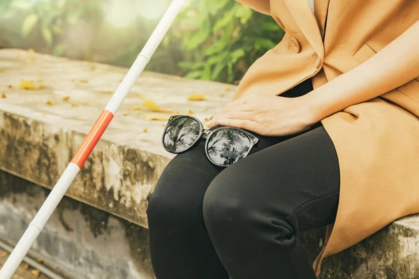 Young blind woman with vision disabilities takes off sunglasses protects eyesight and holds blind cane sitting calmly on concrete platform relaxing in park : Self care blind disabled woman concept.