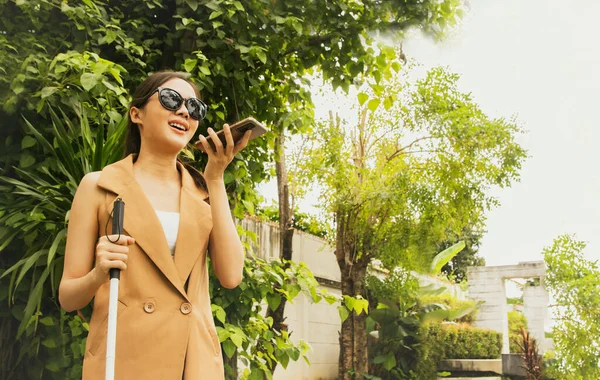 Happy beautiful blind asian woman wearing dark glasses and holding crutches sitting using digital smartphone accessibility application with voice technology in visually impaired assistance features.