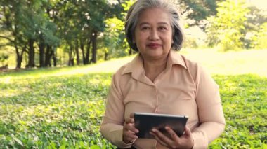 Asian senior woman sitting and chatting with lover, communicating long distance online with tablet with happiness, happy heart, healthy when sit and relax in shady garden suitable for relaxation.