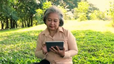 Asian senior woman sitting and chatting with lover, communicating long distance online with tablet with happiness, happy heart, healthy when sit and relax in shady garden suitable for relaxation.