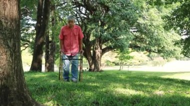 Asian elderly man recovering from paralysis with physiotherapy, using crutches to support his body and walking on the grass, walking slowly to safety.