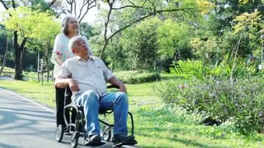 Elderly couple health care relaxing in the garden, with the wife in a wheelchair walking slowly and enjoying the view of the garden with the disabled husband in the morning.
