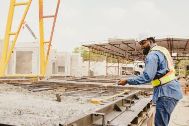 African American male lead architect diligently uses builder's tool to spread cement poured into a solidly defined structural block for prefabrication in an outdoor construction yard. clipart