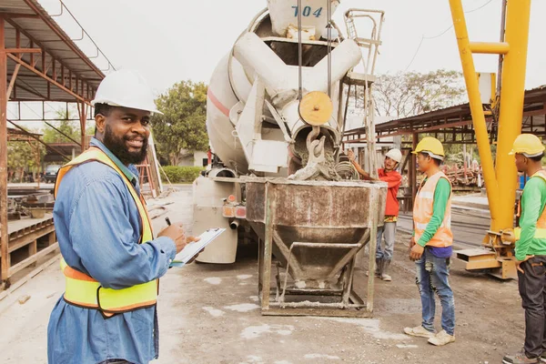 African American foreman inspects the pouring of mortar from a ready-mixed cement truck by team of men using pocket concrete tools or equipment for precast house wall construction.