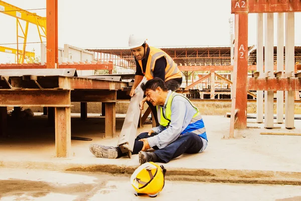 Industrial plant accident large steel bar from high platform falls senior male worker\'s leg prefabricated house wall production site with safety worker helping lift the steel from his painful ankle.