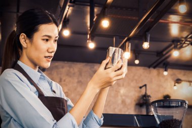 Kahve dükkanındaki iş: Güzel Asyalı barista titizlikle bir sürahiye bakıyor ve müşteriler için güzel bir sütlü kahve menüsünü süslemek için bir sürahi süt tutuyor..