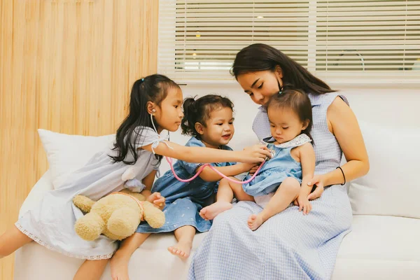 Happy single mother raising three daughters at home : Mother takes care of her daughter sitting together friendly having fun as a doctor and holding stethoscope her youngest daughter in a cozy home.