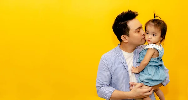 Father\'s happiness : Handsome father standing in the yellow studio holding cute and mischievous youngest daughter kisses cheeks with love Caring for the playful innocence of toddler girls.