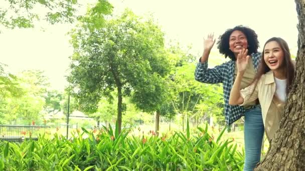 言ってやるがいい アジア系アメリカ人とアフリカ系アメリカ人の親友2人が公園で遊んでいて 大きな木の陰で密かに立って ユーモラスな笑顔で挨拶している — ストック動画