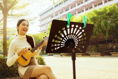 Güzel Asyalı öğretmen oturur ve okul binasının önünde ukulele çalar. Öğrencilere müzik dersi vermeye hazırlanır. Şarkı söyler, oturur ve keyifle notalara bakar..