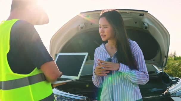 男性の整備士は 旅行中に彼女の車を墜落させた未知の美しいアジアの女性の運転によって引き起こされる損傷を相談し 評価するエンジンを検査するために立っています — ストック動画
