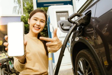 Sıradan pozlarda gülümseyen genç bir kadın, reklam için beyaz ekranlı cep telefonunu gösteriyor. Benzin istasyonlarında çalışan elektrikli araçları ayırtmak için kullanılan bir uygulama tanıtıyor..