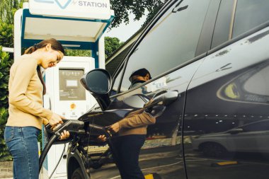 Asyalı bir kadın arabasının şarj kablosu tıkaçını tutuyor, karnı rahat servis istasyonundan memnun: Genç bir kadın yolda ekonomik ve maliyetli bir elektrikli araç kullanıyor.