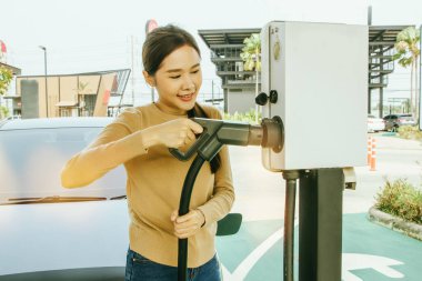 Elektrikli araba kullanan Asyalı bir kadının elinde şarj kablosu olan DC şarj istasyonuna giderken çekilmiş fotoğrafı: Uygun EV elektrikli yakıt istasyonu kadınlara harika bir konsept hissettiriyor.