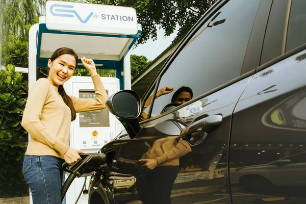 stock image Smiling asian woman raises her hands in delight as the battery of the electric car is nearly full in no time : Woman holding charger plug ready to unplug from crotch of her car.