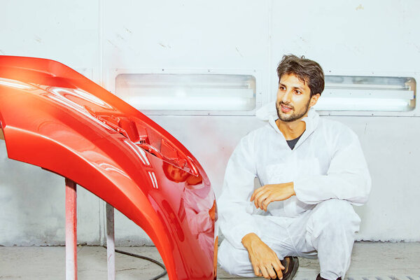 Portrait handsome skilled painter technician professional white uniform sitting and admiring the masterpiece job painted red fiberglass car front bumper successfully repaired in the paint room.