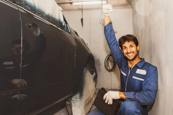 Handsome skilled car painter dressed in automotive painter\'s uniform sits happily holds up his fist admires the new blue paint masterpiece in the paint room smiling and looking into the camera.