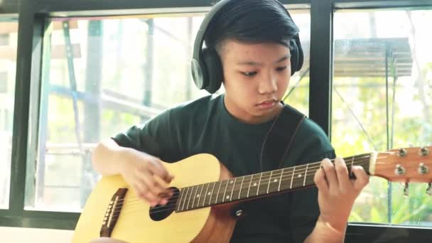 有音乐天赋的年轻的亚洲男孩坐在家里 喜欢在窗边弹吉他 — 图库视频影像