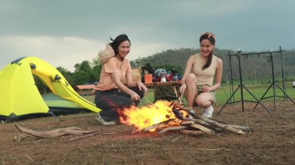 假日露营活动 两名兴奋的亚洲女孩在一座被茂密的森林和高山环绕的野营帐篷前燃起的篝火前 — 图库视频影像