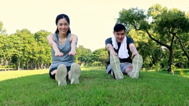 朝の木の下の公園の芝生の中で暖かい座っているアジアの医療カップルは腕を伸ばし 足は筋肉をリラックス運動する前に怪我を防ぐ — ストック動画