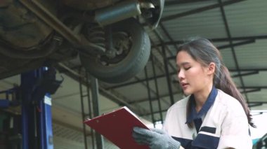 Asyalı kadın teknisyen araba süspansiyonunu, frenleri ve sol arka tekerleği dikkatle kontrol ediyor. Anormal sesleri dikkatle dinleyin ve garajdaki müşteri araba bakımına rapor edin..