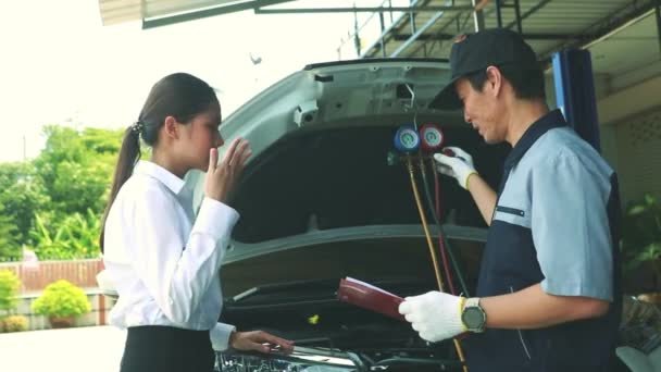男性の自動車整備士は 暑さの中で車を運転しながら損傷や冷媒漏れを修復するために 女性の顧客サービスの車のエアコンシステムを巧みに検査します — ストック動画