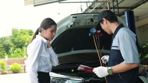 男性の自動車整備士は 暑さの中で車を運転しながら損傷や冷媒漏れを修復するために 女性の顧客サービスの車のエアコンシステムを巧みに検査します — ストック動画