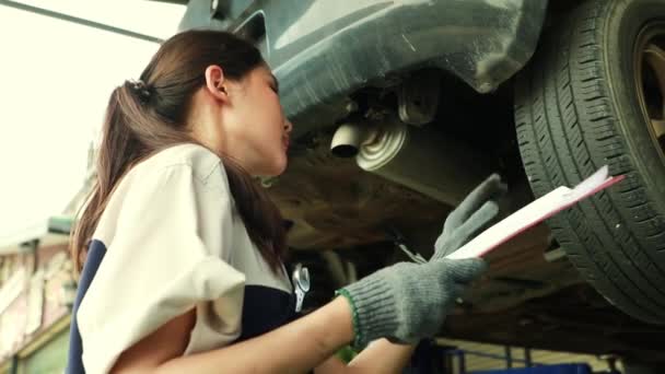 アジアの女性技術者は 車のサスペンションチェックブレーキは慎重に軸受左リアホイールをチェックし ガレージで顧客の車のメンテナンスのためのクリップボードに報告書を記録しました — ストック動画