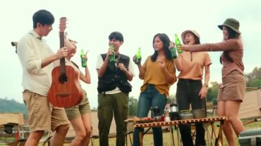 Asyalı erkekler ve kızlar gitar çalıp kadeh kaldırıyor bira şişelerini kırıyor ve mutlu bir şekilde gülüyor. Kamp partisinde yakın dostların iyi niyetine katılın..