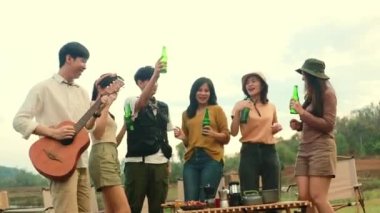 Asyalı erkekler ve kızlar gitar çalıp kadeh kaldırıyor bira şişelerini kırıyor ve mutlu bir şekilde gülüyor. Kamp partisinde yakın dostların iyi niyetine katılın..