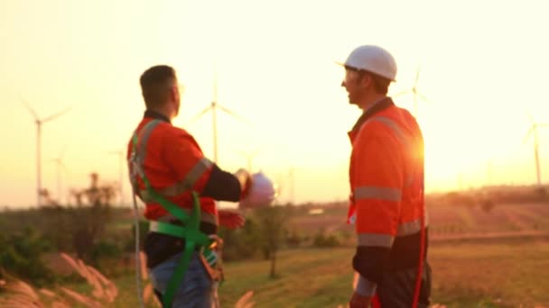 チーム2人の労働者エンジニアの技術者は自然風を使用して日没の作業場の再生可能エネルギー発電所を見ている男性をリラックスさせました 風力タービンは環境のための電気を発生させます — ストック動画