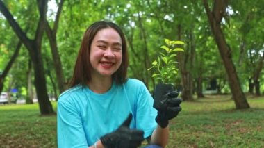 Genç Asyalı kadın bahçedeki çevre için ağaç dikmeye gönüllü oluyor. Yeşillik ekliyor, elinde mutlu bir gülümsemeyle genç ağaç fidanını kameraya bakarken gösteriyor..