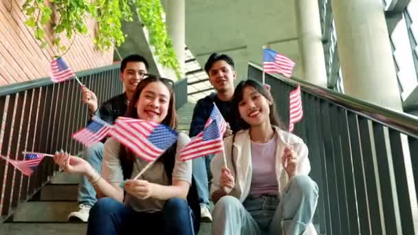 7月4日を祝うアメリカの国旗を保持し 振るグループアジアの大学生は 学校の建物の階段に座っています — ストック動画