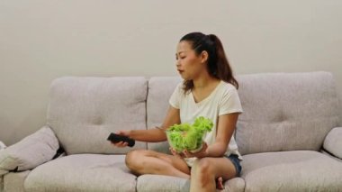 Neşeli Asyalı kadın, TV 'de eğlence şovu filmleri izlerken kanepede yeşil salata yiyor: Sağlık hizmetleri, yağ diyeti ve kilo verme kavramını azaltmak.